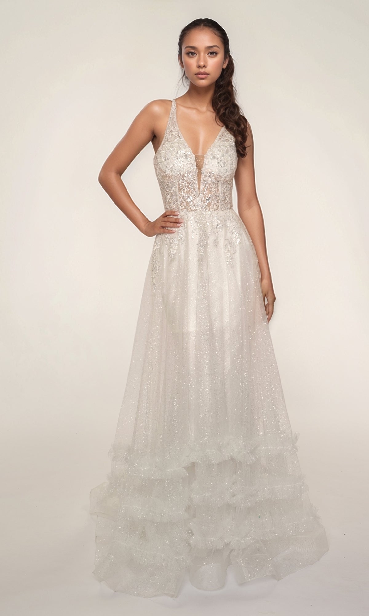 A woman models the Nina Canacci 7607, a sleeveless, V-neck, floor-length white formal dress with a fitted silhouette, sheer lace bodice, and tiered ruffle skirt, standing against a plain light background.